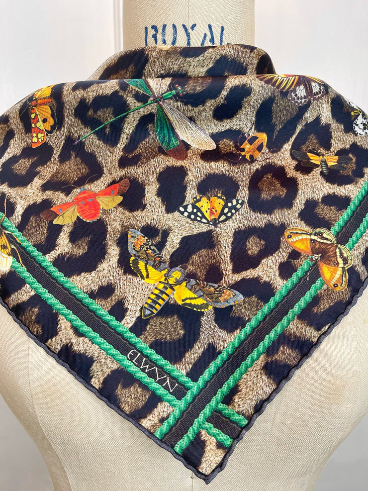 a closeup of a designer silk bandana. The Italian silk bandana Features a Leopard fur print, adorned with multicolored entomological moths, butterflies, beetles, and dragonflies and framed by a rope ribbon border. Imagined to be a sleeping Leopard, where butterflies and the such come to land and rest on her back in a daydream... This is a hand sewn silk scarf.