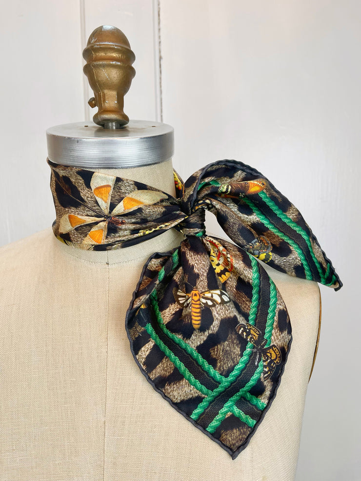 A designer silk bandana is tied around the neck of a form like a side bow. The Italian silk bandana Features a Leopard fur print, adorned with multicolored entomological moths, butterflies, beetles, and dragonflies and framed by a rope ribbon border. Imagined to be a sleeping Leopard, where butterflies and the such come to land and rest on her back in a daydream... This is a hand sewn silk scarf.