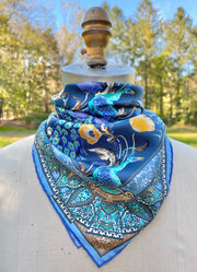 An Italian silk bandana tied around a form with a wallpaper type print, featuring radiant peacocks, amidst sprigs of yellow poppies, surrounded by ornate ribbons folding around the border.