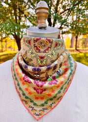 An Italian silk bandana featuring a geometric design, mixing earth tones and pops of brightly colored etymological butterflies tied around a mannequin's neck in the woods. 