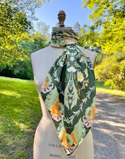 This bespoke, designer silk Scarf is Art Nouveau inspired featuring a kaleidoscope of fantastical flowers, butterflies and cockatoos rising from the curling foliage. The scarf is tied around a form in the woods.