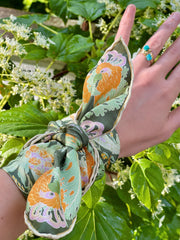This bespoke, luxury silk bandana is Art Nouveau inspired featuring a kaleidoscope of fantastical flowers, butterflies and cockatoos rising from the curling foliage tied around a model's wrist with a scenic green background.