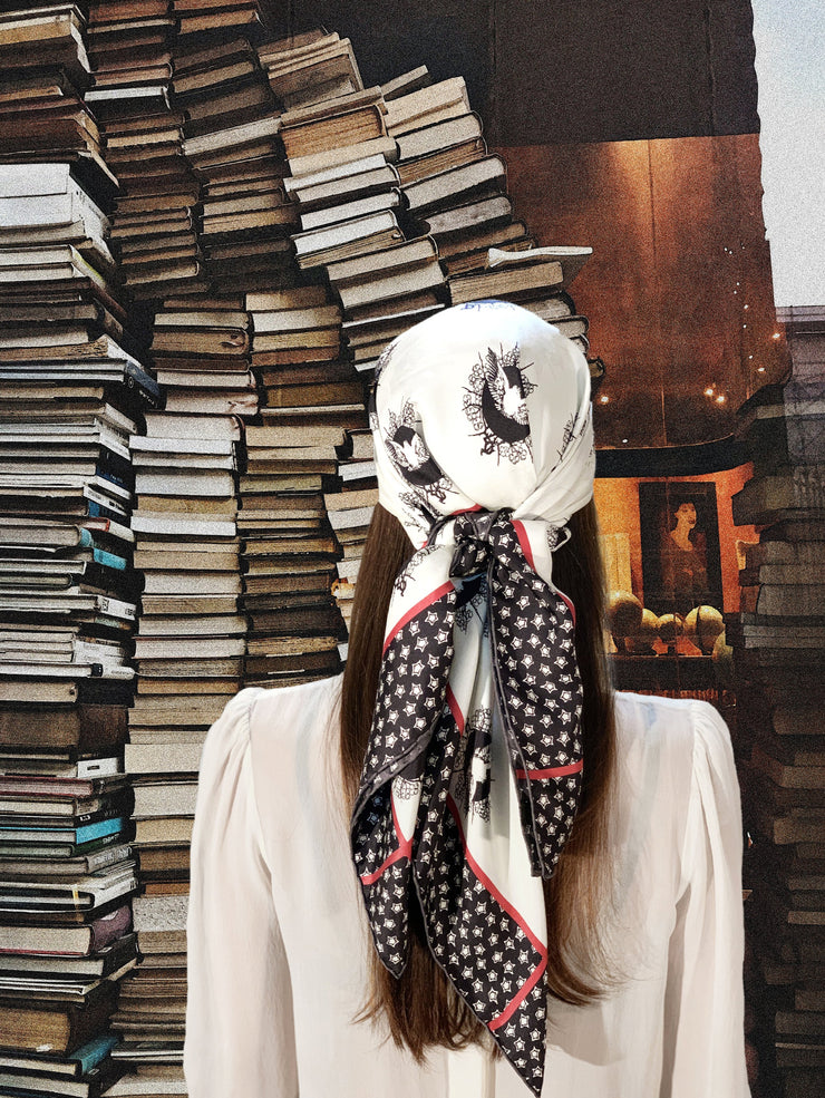 classic female standing in front of a fantastical storefront window piled with books, wearing a luxury, bespoke Elwyn New York silk scarf on her head with black and white, whimsical, polka dot, bird and stars, storybook print