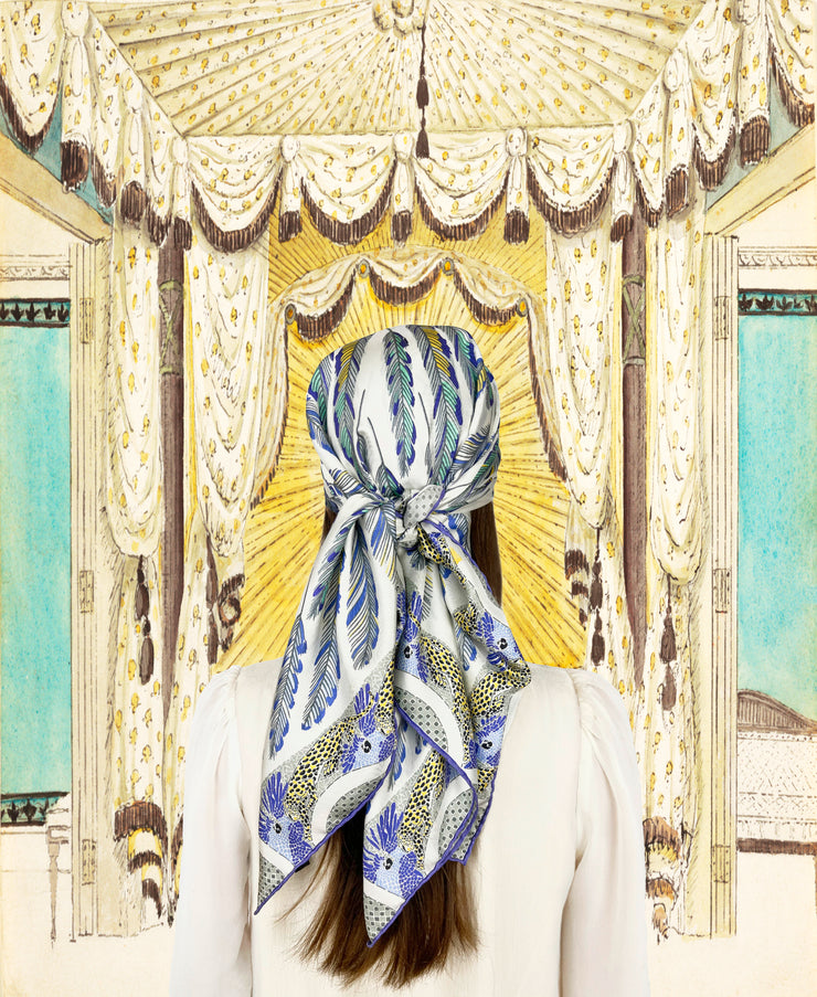 Fantasy illustration of a back view of woman wearing a bespoke Elwyn New York silk scarf on her head with a delightful design of swirling feathers, surrounded by creeping jaguars and parrots in the graphic border.