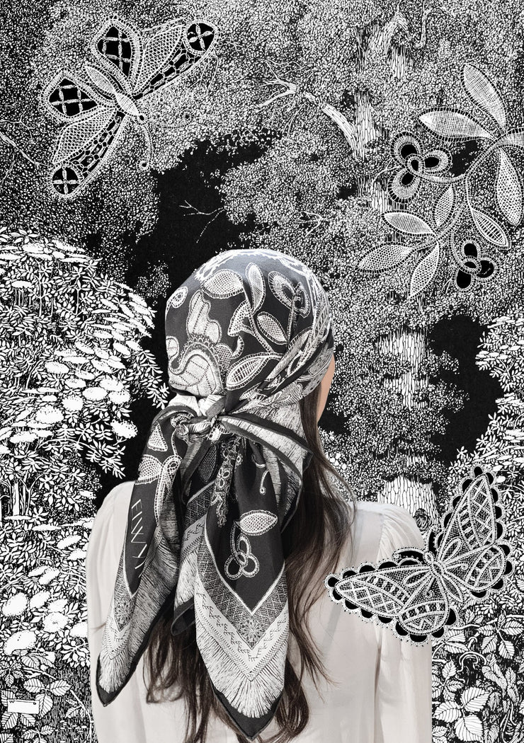 classic female traveler standing in an illustrated blooming garden, wearing bespoke, luxury, black and white Elwyn New York silk scarf on her head with vintage lace print and graphic fringe border