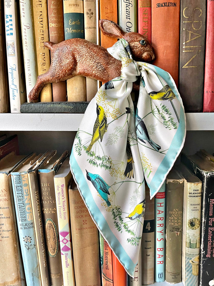 Closeup of a bespoke Elwyn New York silk bandana tied around the neck of a rabbit figurine with a Charming blue and yellow warbler birds, flying and perched amidst delicate wild flowers. Inspired by nature found in the North East.