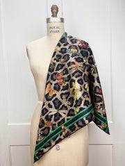 A designer silk scarf draped on one shoulder of a form.  The Italian silk scarf designed in New York, Features a Leopard fur print, adorned with multicolored entomological moths, butterflies, beetles, and dragonflies and framed by a rope ribbon border.  Imagined to be a sleeping Leopard, where butterflies and the such come to land and rest on her back in a daydream...  