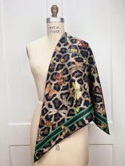 A designer silk scarf draped on one shoulder of a form.  The Italian silk scarf Features a Leopard fur print, adorned with multicolored entomological moths, butterflies, beetles, and dragonflies and framed by a rope ribbon border.  Imagined to be a sleeping Leopard, where butterflies and the such come to land and rest on her back in a daydream... This is a hand sewn silk scarf 