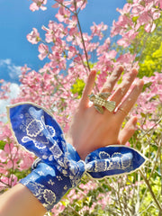 a closeup of a bespoke Elwyn New York bandana tied around the wrist of a model like a bib with an ornate, blue and white, vintage-pastoral bandana design. Classic, feminine, and romantic.