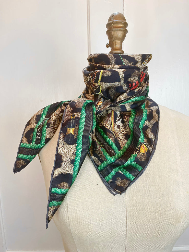 A designer silk scarf Tied around the neck of a form.  The Italian silk scarf Features a Leopard fur print, adorned with multicolored entomological moths, butterflies, beetles, and dragonflies and framed by a rope ribbon border.  Imagined to be a sleeping Leopard, where butterflies and the such come to land and rest on her back in a daydream...  