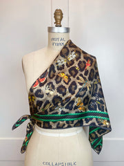 A designer silk scarf tied like a blouse on a form.  The Italian silk scarf Features a Leopard fur print, adorned with multicolored entomological moths, butterflies, beetles, and dragonflies and framed by a rope ribbon border.  Imagined to be a sleeping Leopard, where butterflies and the such come to land and rest on her back in a daydream...  
