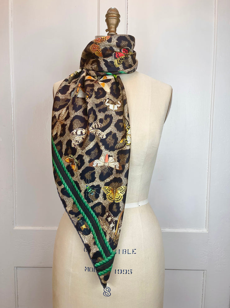 A designer silk scarf wrapped around the neck of a form.  The Italian silk scarf Features a Leopard fur print, adorned with multicolored entomological moths, butterflies, beetles, and dragonflies and framed by a rope ribbon border.  Imagined to be a sleeping Leopard, where butterflies and the such come to land and rest on her back in a daydream...  