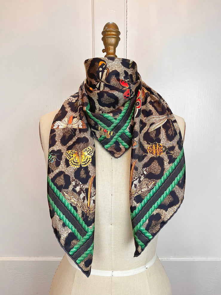 A designer silk scarf wrapped around the neck of a form.  The hand sewn silk scarf Features a Leopard fur print, adorned with multicolored entomological moths, butterflies, beetles, and dragonflies and framed by a rope ribbon border.  Imagined to be a sleeping Leopard, where butterflies and the such come to land and rest on her back in a daydream...  