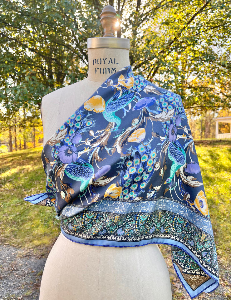 An Italian silk scarf designed in New York with a wallpaper type print, featuring radiant peacocks, amidst sprigs of yellow poppies, surrounded by ornate ribbons folding around the border tied draped over  a dress form's shoulder.