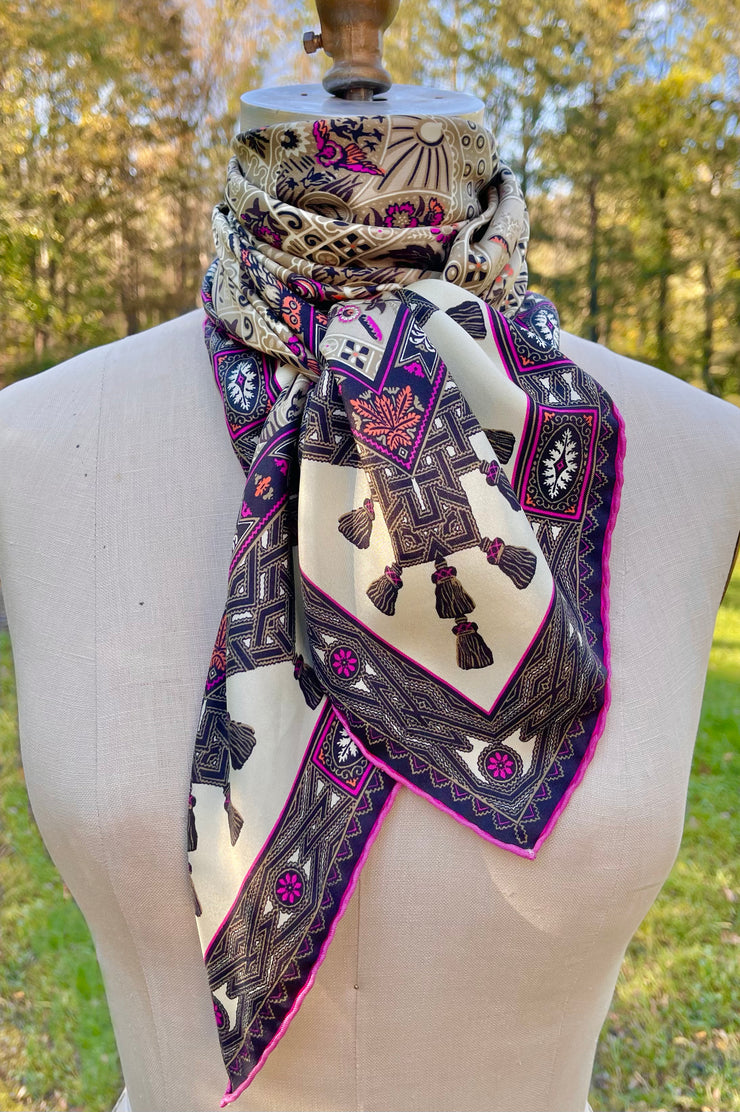 An Italian silk scarf featuring a Anglo-Japanesque print with intersecting geometric forms, framing little garden, owl and cat motifs, surrounded by a woven ribboned border and dangling tassels with pops of fuchsia and orange throughout. 