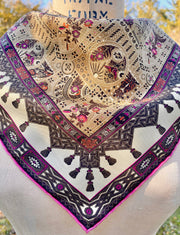 An Italian silk bandana featuring an Anglo-Japanesque print with intersecting geometric forms, framing little garden, owl and cat motifs, surrounded by a woven ribboned border and dangling tassels with pops of fuchsia and orange throughout tied around the neck of a form.