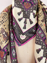 A dress form Italian silk scarf around the neck with an Anglo-Japanesque print with intersecting geometric forms, framing little garden, owl and cat motifs, surrounded by a woven ribboned border and dangling tassels with pops of fuchsia and orange throughout.