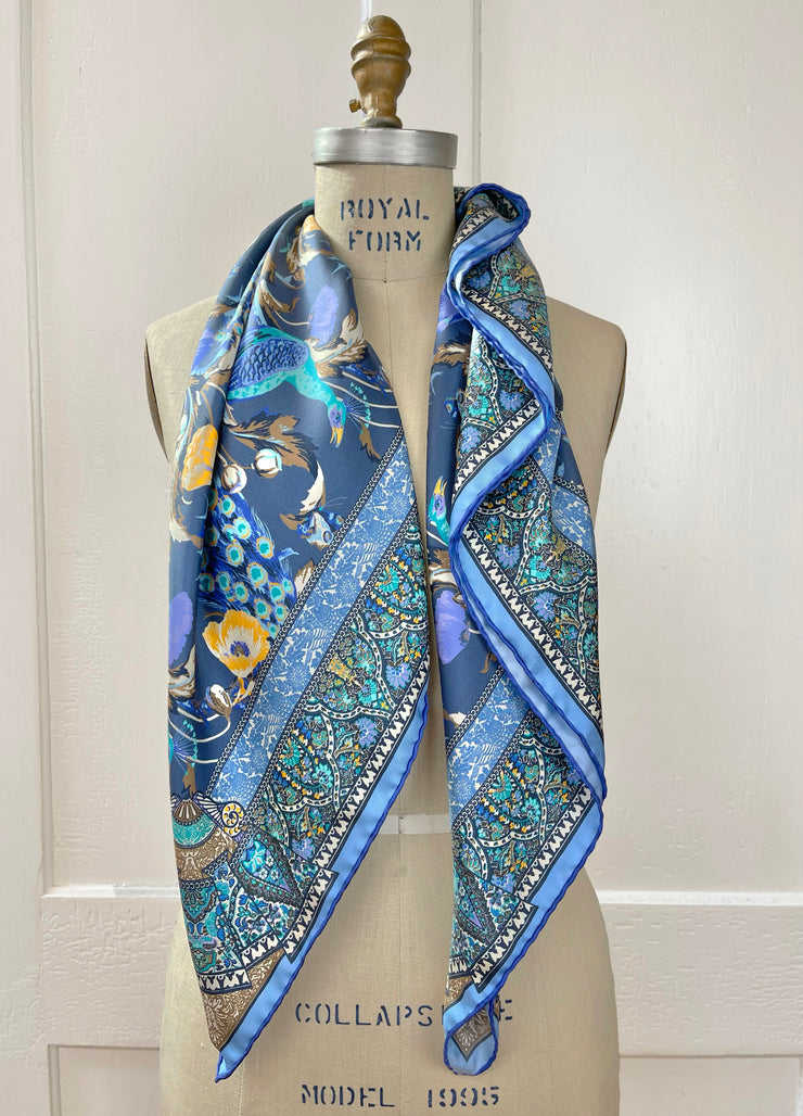 An Italian silk scarf designed in New York with a wallpaper type print, featuring radiant peacocks, amidst sprigs of yellow poppies, surrounded by ornate ribbons folding around the border tied around a dress form's neck. 