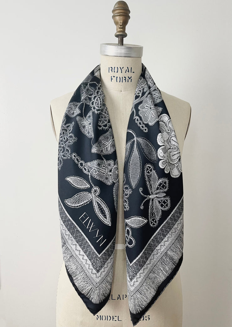 A bespoke, luxury, black and white Elwyn New York silk scarf hanging around the neck of a form with a vintage lace print and graphic fringe border