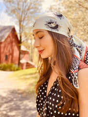 classic woman standing in front a red barn and flowering tree, wearing a luxury, bespoke Elwyn New York silk scarf on her head with black and white, whimsical, polka dot, bird and stars, storybook print