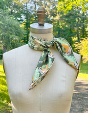 This bespoke, luxury silk bandana is Art Nouveau inspired featuring a kaleidoscope of fantastical flowers, butterflies and cockatoos rising from the curling foliage tied around the neck of a mannequin with a scenic green background.