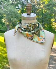 This bespoke, luxury silk bandana is Art Nouveau inspired featuring a kaleidoscope of fantastical flowers, butterflies and cockatoos rising from the curling foliage tied around the neck of a mannequin with a scenic green background.