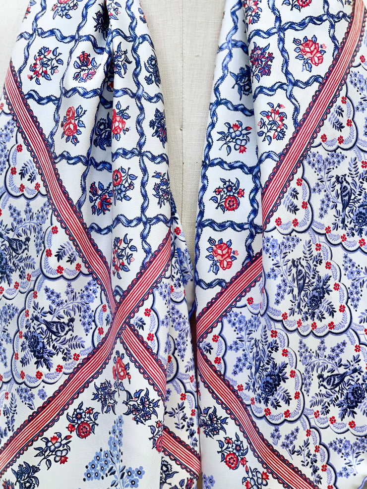 A closeup of the silk scarf. The main pattern is made up of a red, white and blue diamond lattice of curving ribbons, enclosing sprays of flowers and is surrounded by an eyelet lace border, adorned with charming little birds.