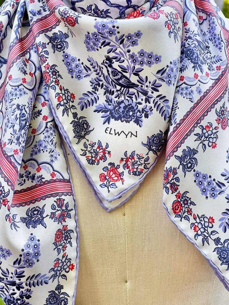 A closeup of the silk scarf. The main pattern is made up of a red, white and blue diamond lattice of curving ribbons, enclosing sprays of flowers and is surrounded by an eyelet lace border, adorned with charming little birds.