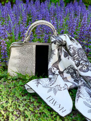 luxury, bespoke Elwyn New York silk scarf draped around her neck with black and white, art nouveau, whimsical, storybook print tied around a vintage handbag set in front a patch of purple wildflowers 