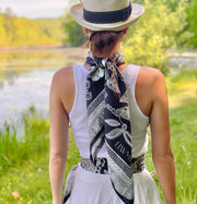 back view of a classic female looking at a pond, wearing bespoke, luxury, black and white Elwyn New York silk scarf around her neck but tied to the back and one tied as a belt.  Both have a vintage lace print and graphic fringe border