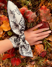 Elwyn New York luxury, bespoke Elwyn New York silk bandana tied around the wrist like a bow with black and white, art nouveau, whimsical, storybook print. Lady's hand resting on a bed of red fall leaves