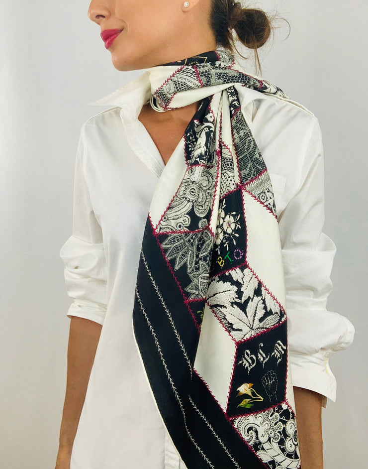 Classic woman wearing bespoke, luxury Elwyn New York silk scarf draped long and loose around her neck. This geometric crazy quilt print is a vintage-modern depiction of the year 2020 filled with digital embroidery and lace of years past.  One can see the words LGBTQ and BLM depicted in this photo