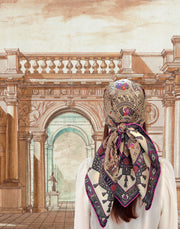 A classic model wearing an Italian silk scarf around her head with an Anglo-Japanesque print with intersecting geometric forms, framing little garden, owl and cat motifs, surrounded by a woven ribboned border and dangling tassels with pops of fuchsia and orange throughout.