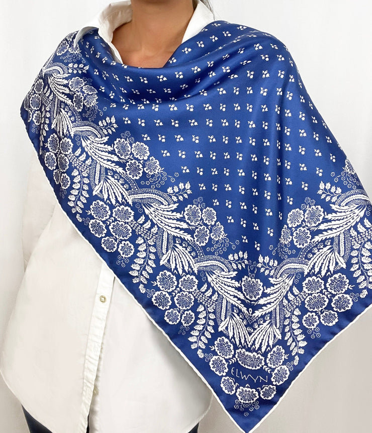 Woman wearing a personalized bespoke Elwyn New York Scarf draped  draped on one shoulder with an ornate, blue and white, vintage-pastoral bandana design. Classic, feminine, and romantic