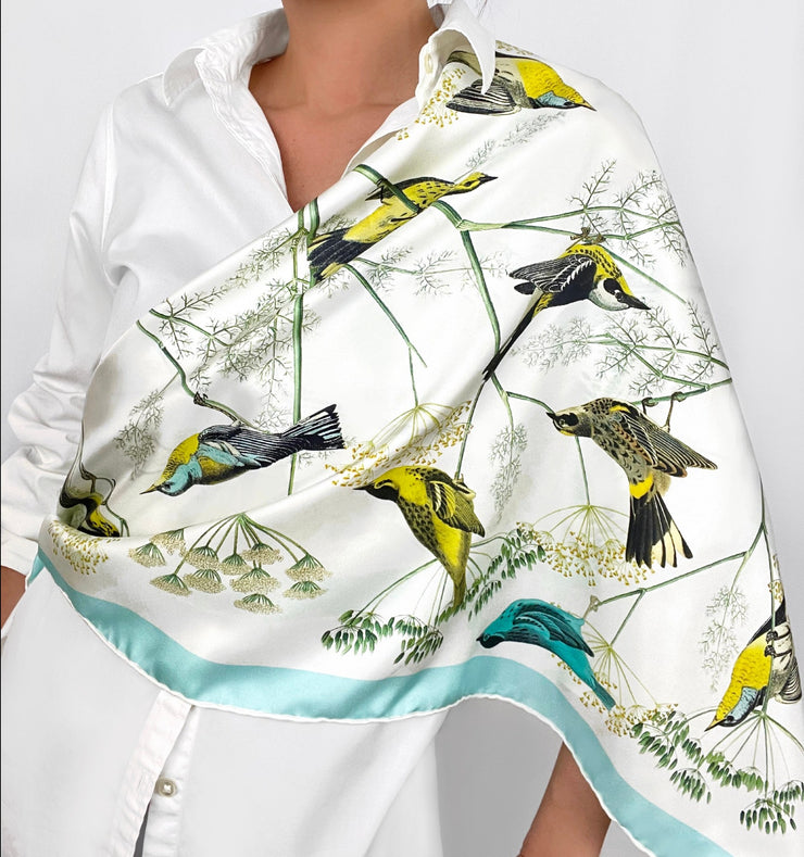 a classic woman wearing a bespoke Elwyn New York silk scarf draped around one shoulder with a Charming blue and yellow warbler birds, flying and perched amidst delicate wild flowers. Inspired by nature found in the North East.