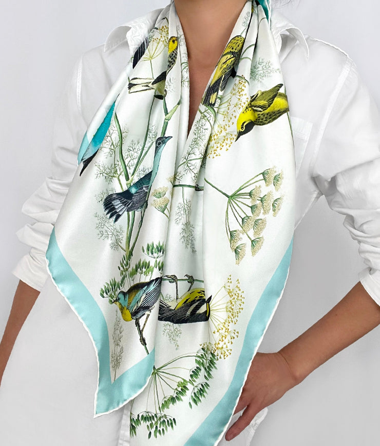 a classic woman wearing a bespoke Elwyn New York silk scarf draped around her neck with a Charming blue and yellow warbler birds, flying and perched amidst delicate wild flowers. Inspired by nature found in the North East.